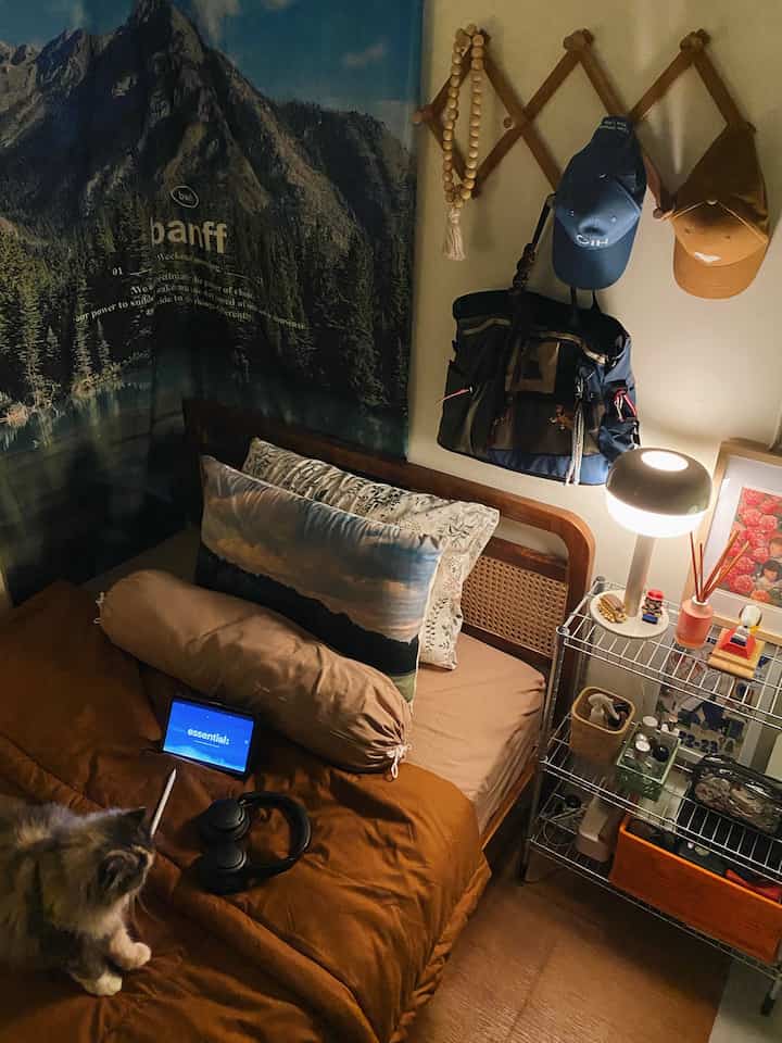 Natural color bedroom featuring brown bedspread and wooden bed frame, wall hooks with bags and hats, cozy space with lamp on metal shelf