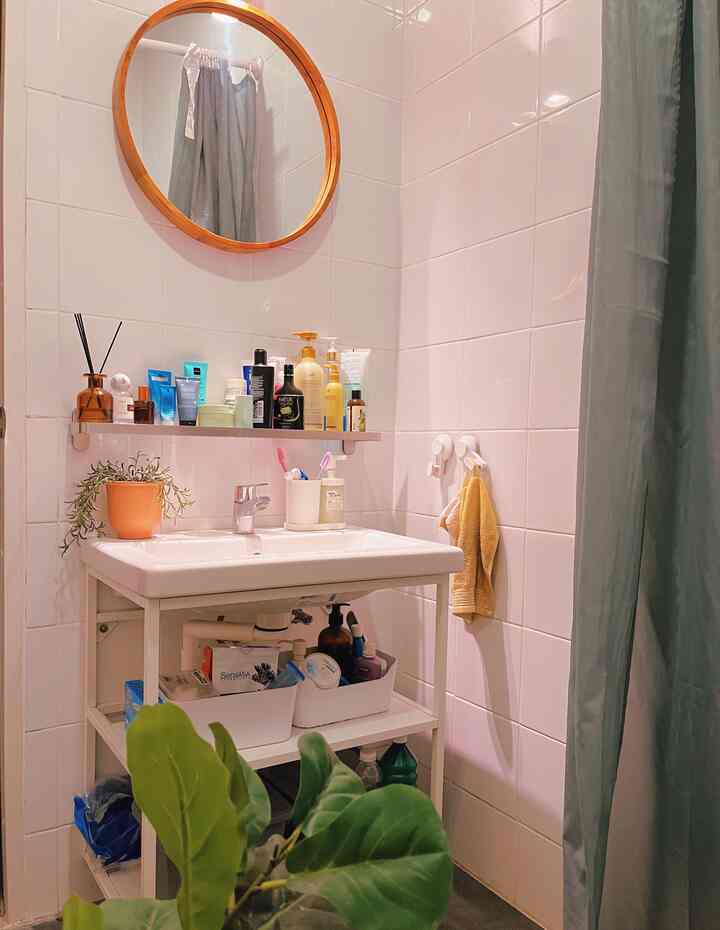 White tiled bathroom featuring a round wooden mirror, organized sink shelf with toiletries, and a clean, natural ambiance