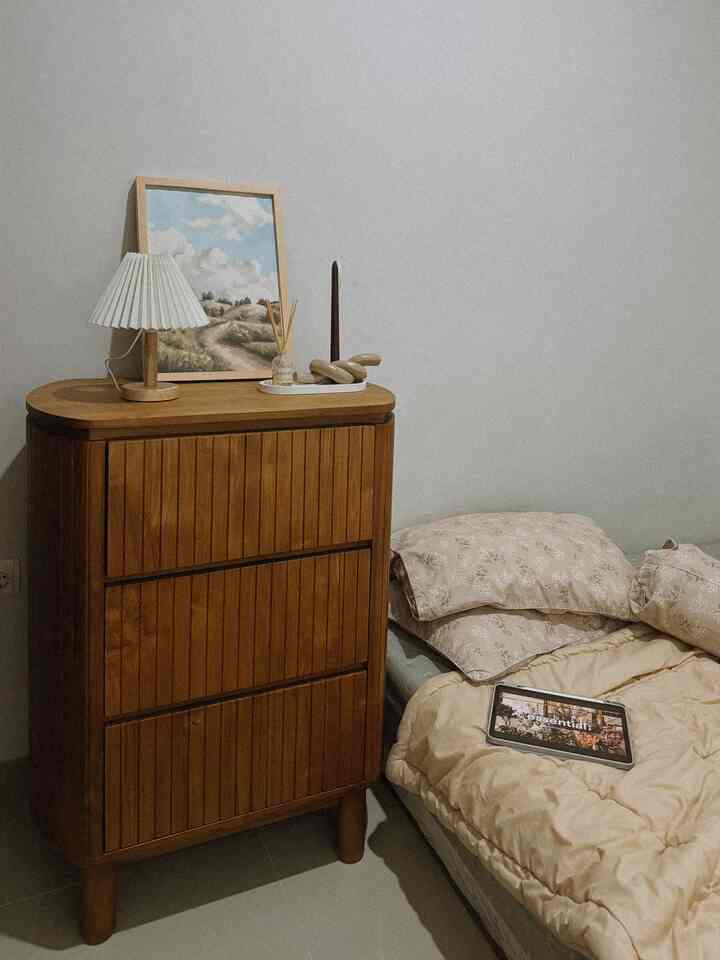 Natural wood-toned bedroom featuring a wooden chest of drawers, bedspread in beige tones, and decor elements including table lamp, painting, diffuser, and candle creating a calm atmosphere