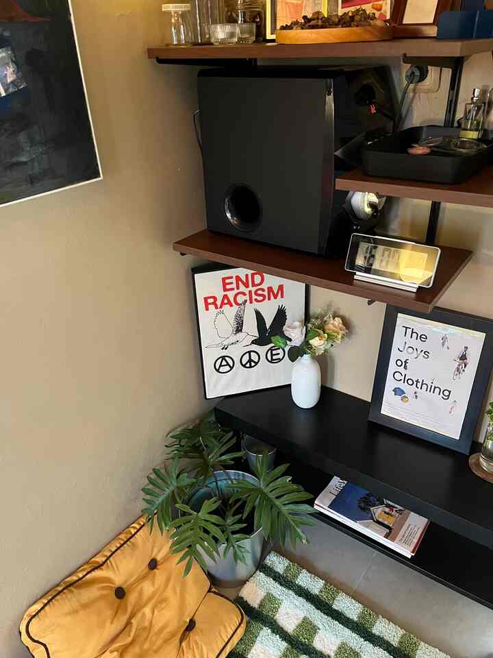 Natural tone wall corner space featuring black corner shelving, yellow cushion, and large green plant creating a cozy atmosphere