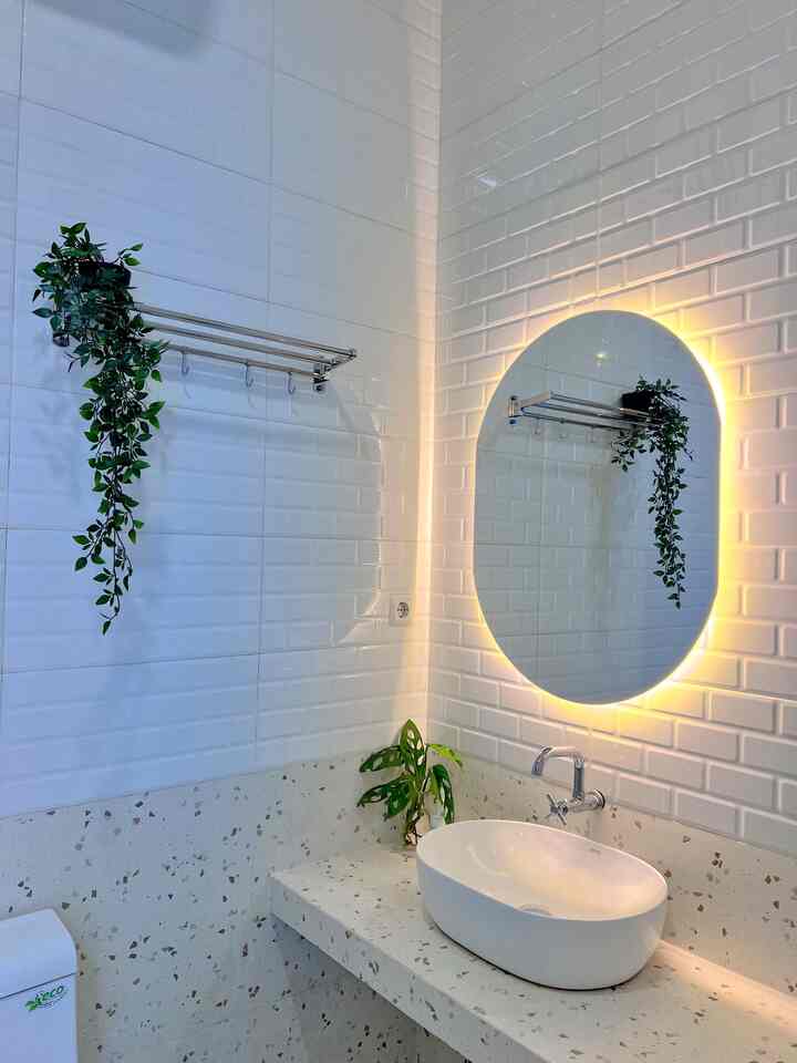 Bathroom with white tiled walls and terrazzo countertop featuring a white basin sink and LED backlit oval mirror, accented by green plants