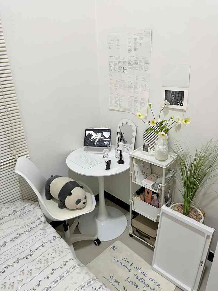 A white-toned small bedroom corner featuring a round desk, office chair, storage rack, and floral decor creating a cozy workspace