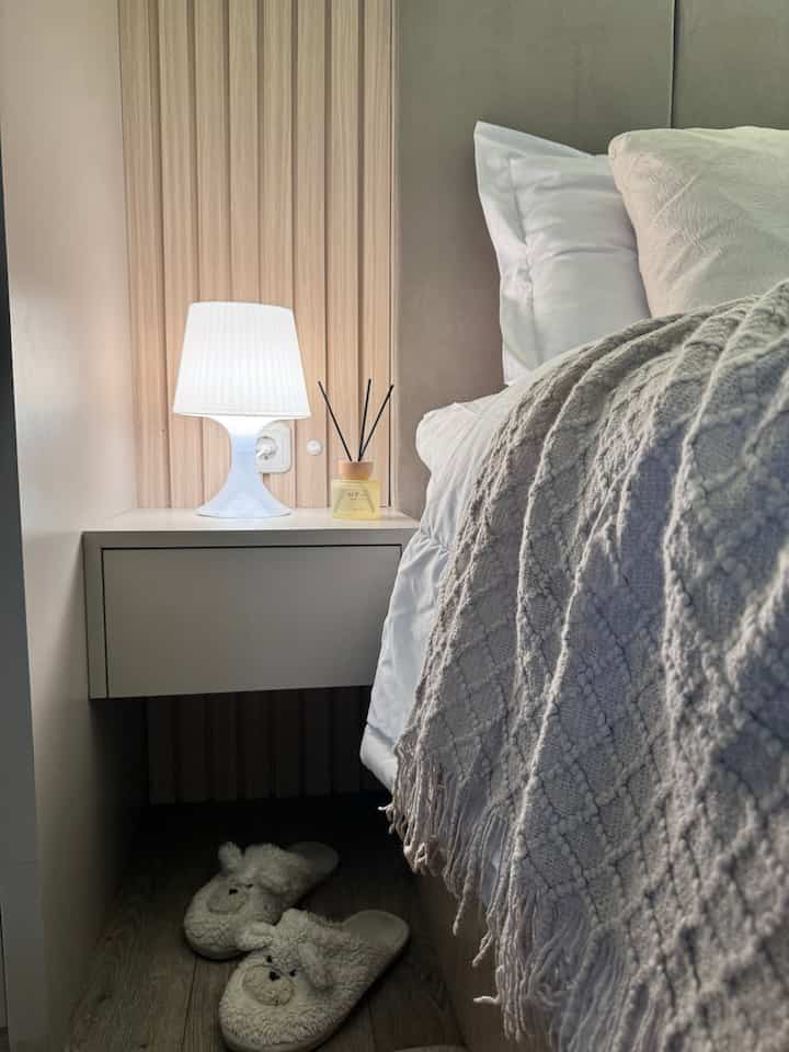 Natural toned bedroom corner featuring a white nightstand with table lamp and diffuser, cozy bed blanket and plush slippers beside bed in narrow space