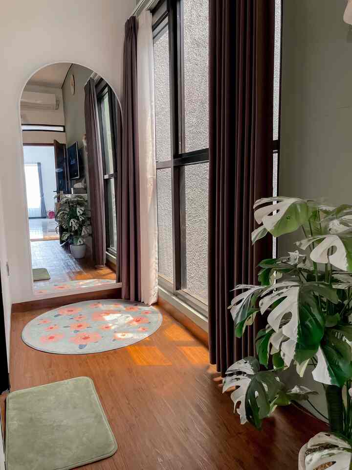 A warm natural-toned narrow bedroom corner featuring a large full-length mirror, round carpet, and artificial Monstera plant creating a cozy interior