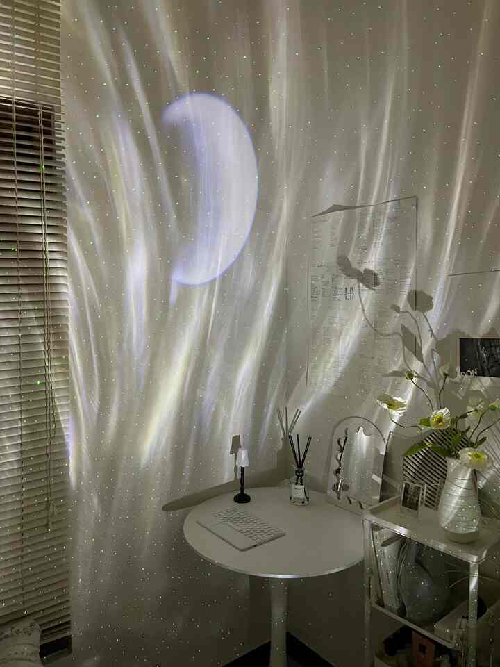 A small bedroom in white and beige tones featuring a round table, flowers, and galaxy projector lights creating a cozy, dreamy atmosphere
