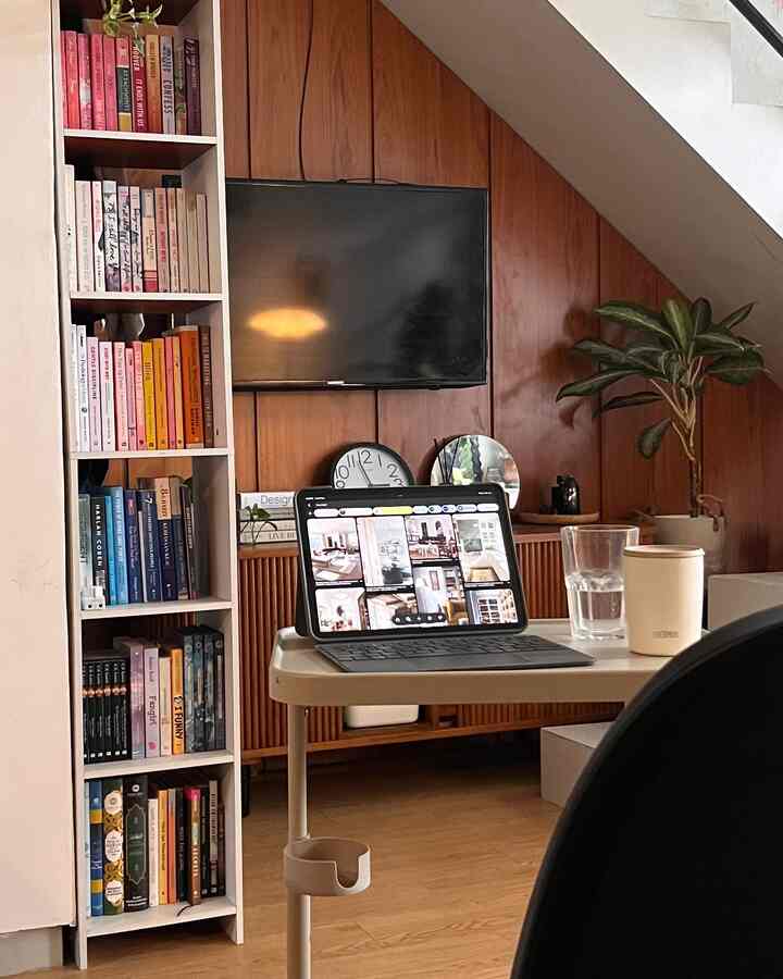 Warm wood-tone wall with bookshelf forms a small home office space, featuring a desk with tablet and plant creating cozy atmosphere