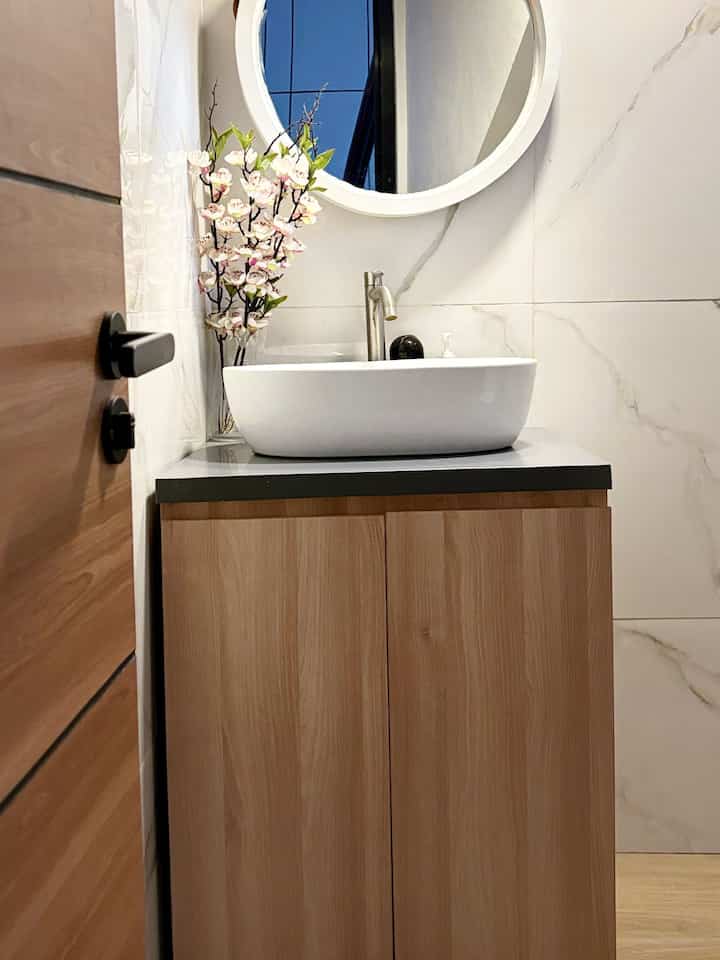 Light brown and white toned powder room featuring a round mirror, ceramic sink, and wooden vanity creating a natural atmosphere