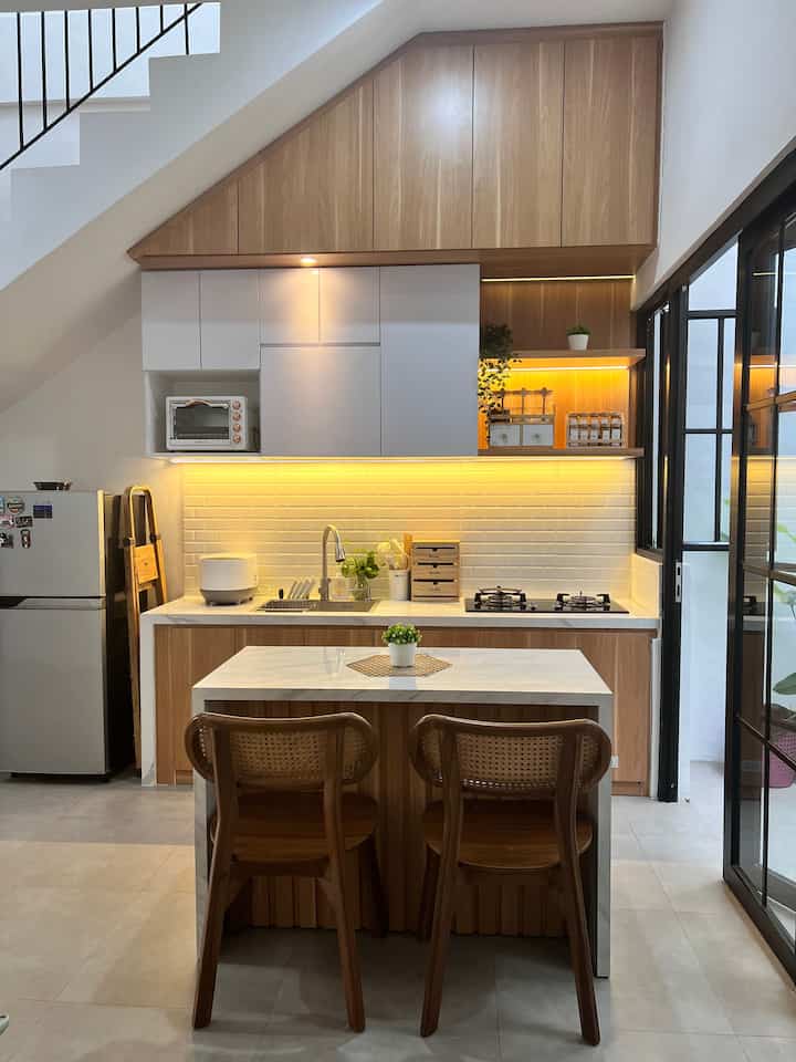 Compact kitchen under stairs featuring wood tone cabinets and white backsplash with warm lighting and efficient storage