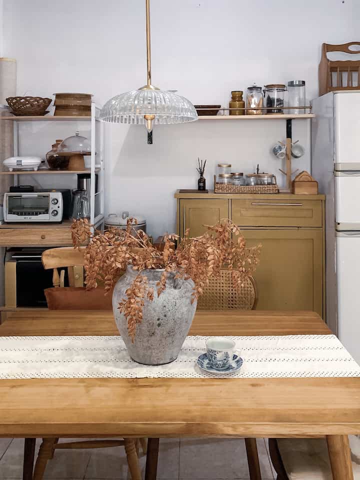Beige and natural toned kitchen space featuring wooden dining table, chairs, pendant light, and assorted kitchenware in cozy arrangement