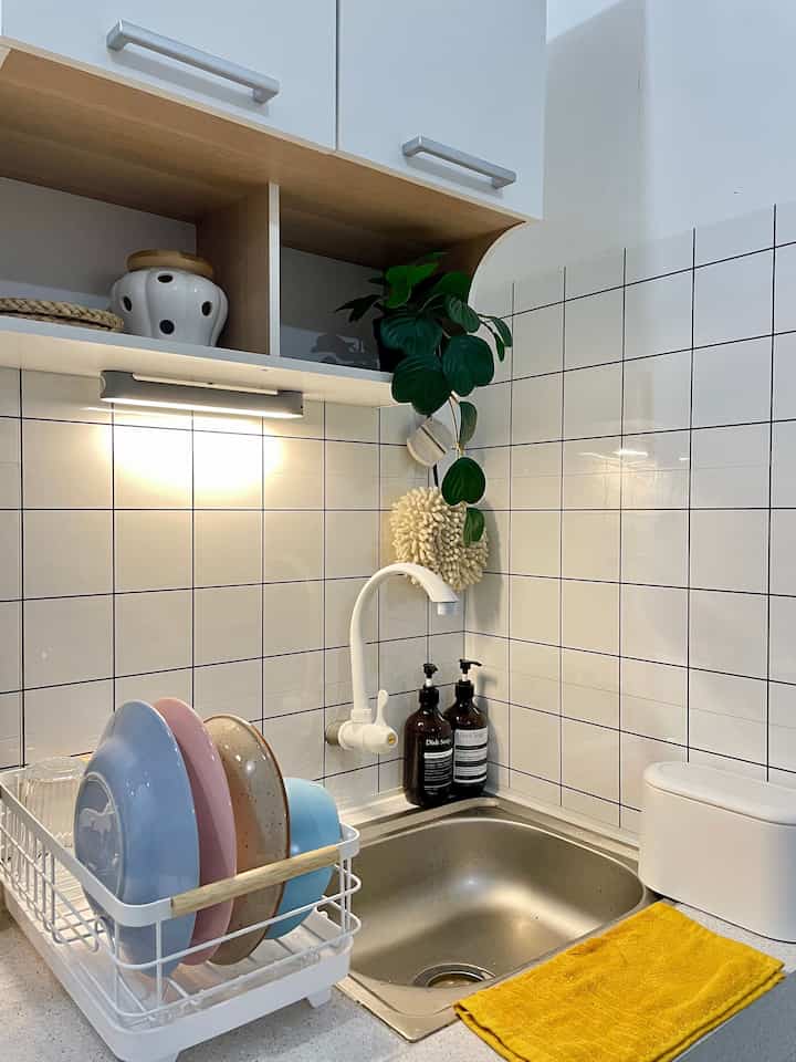 Compact kitchen space with white tiled walls and sink area featuring a white dish rack and yellow towel