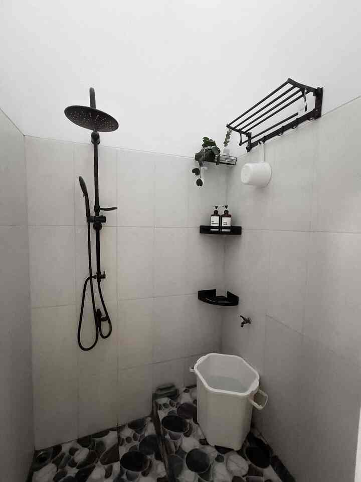 Narrow, white-toned bathroom with black shower fixture, wall shelves, and a white bucket, featuring a minimalist design