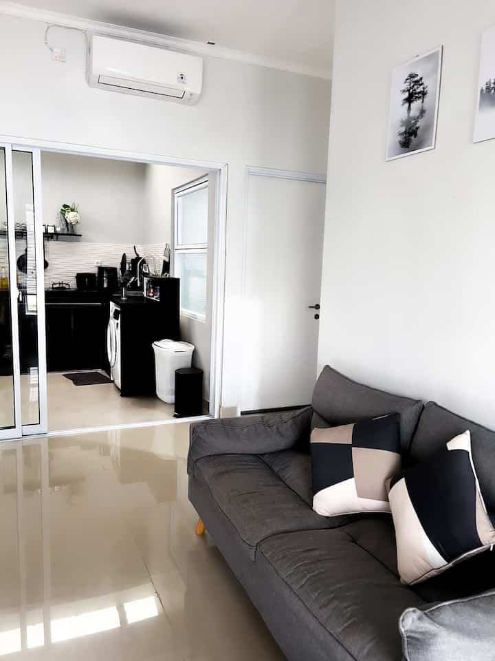 A modern living room in gray and white tones, featuring a gray sofa centrally and a kitchen visible through glass sliding doors