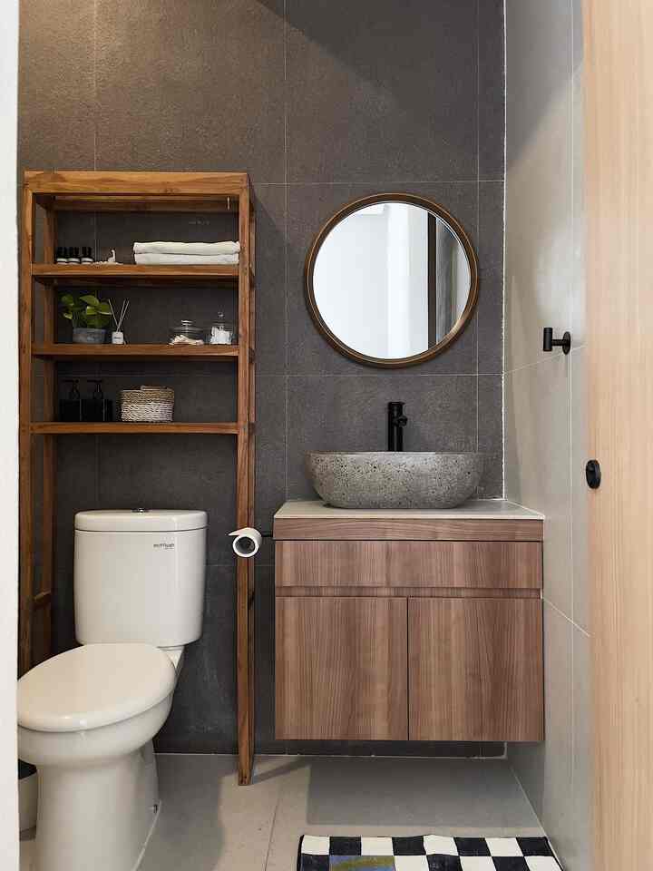 Compact bathroom featuring gray tiled walls, brown wooden vanity and shelves with round mirror and stone wash basin