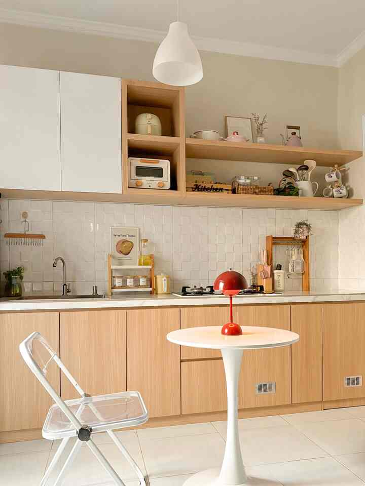 A simple, compact single-wall kitchen with white tiled backsplash and wood-tone cabinets, featuring a white round table and transparent folding chair