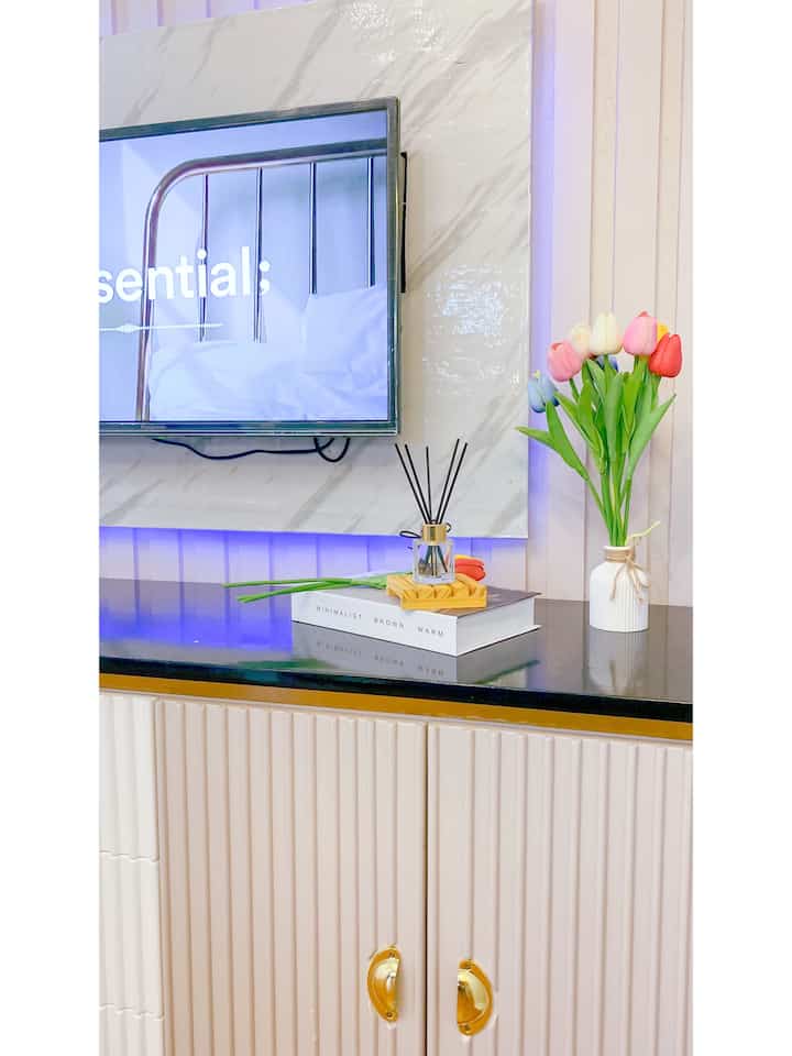 White and gold toned living room featuring TV stand, marble patterned wall panel, and colorful tulip flower arrangement