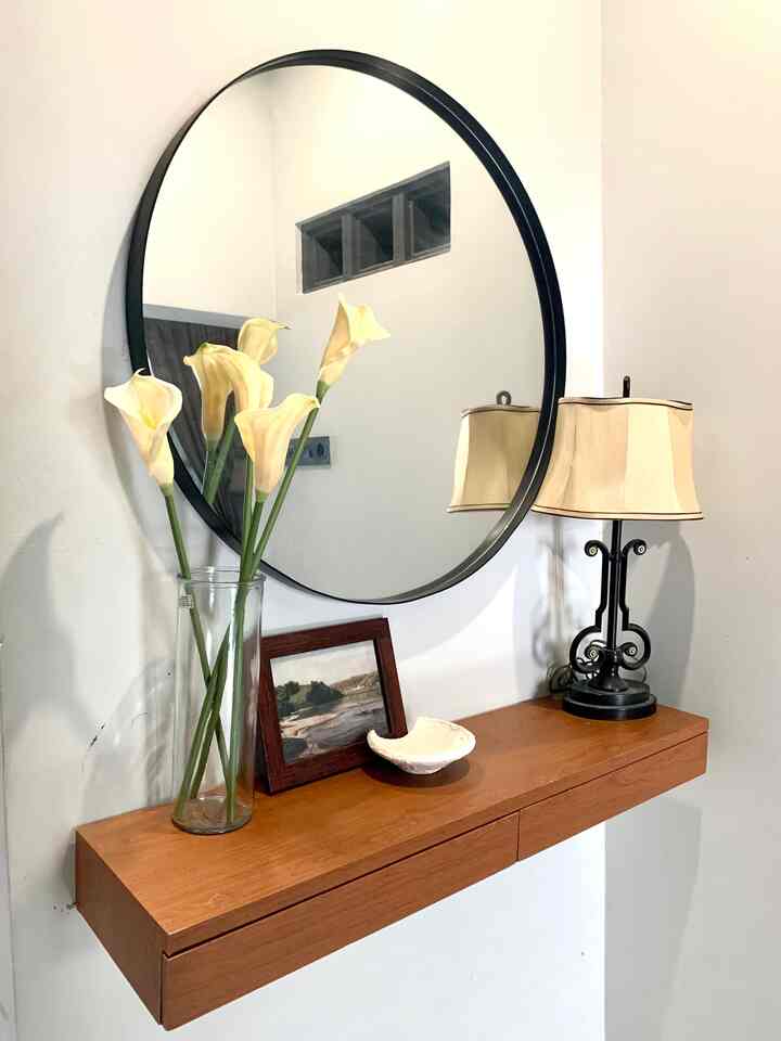 Natural color space featuring a wooden floating shelf with drawers, round mirror, vase with flowers, and a table lamp creating a cozy ambiance