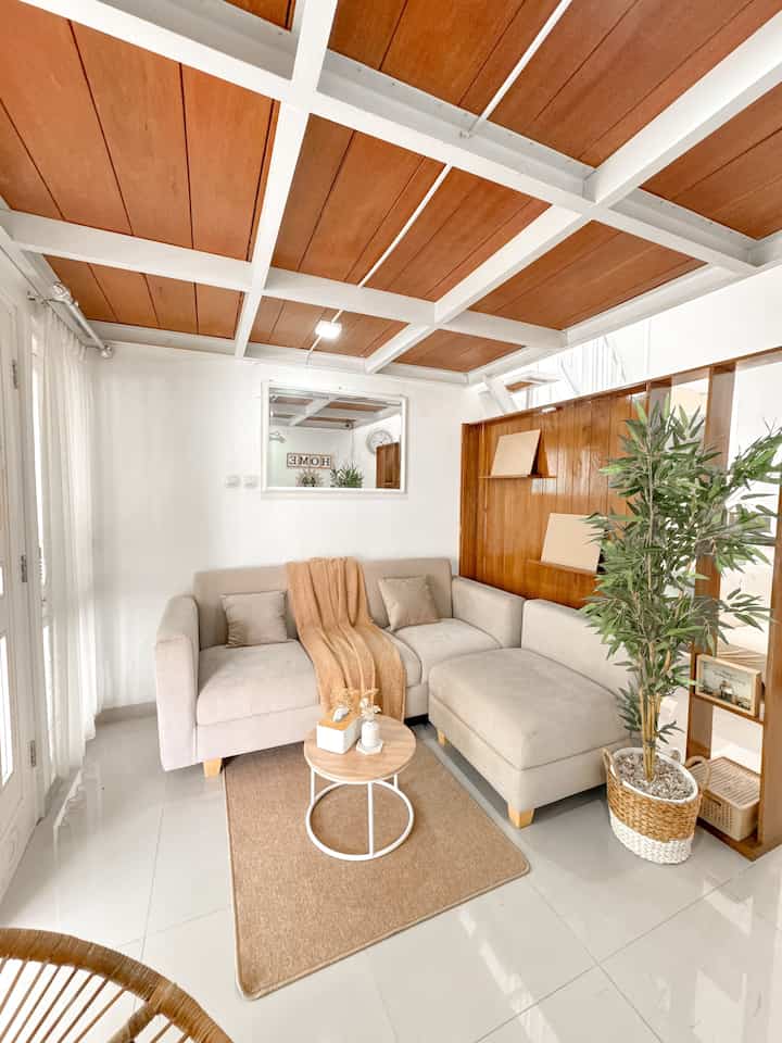 A warm-toned living room with wood tone and white elements, featuring an L-shaped sectional sofa, round coffee table, and artificial bamboo plant