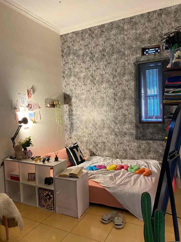 Compact studio bedroom with pink bedsheet, white walls, shelving unit, and blinds on window