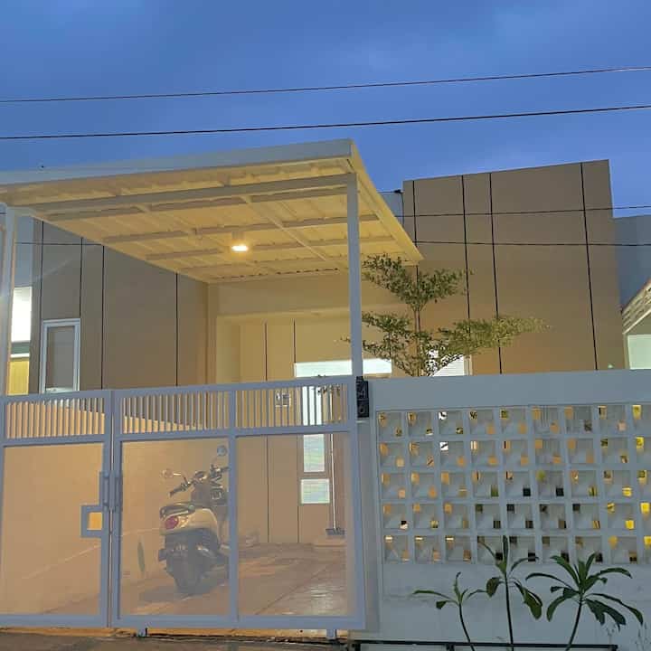 Rumah ini memiliki desain minimalis, dengan dominasi warna putih pada eksteriornya. Pagar depan dengan motif sederhana yang memberi kesan terbuka namun tetap menjaga privasi. Di bagian depan, terdapat sebuah carport yang cukup luas, dilengkapi dengan atap datar yang serasi dengan desain rumah. Carport ini cukup untuk menampung satu motor atau mobil kecil.

Dinding rumah berwarna krem dengan aksen garis-garis vertikal hitam, memberikan sentuhan geometris dan kontemporer pada fasadnya. Di samping pagar, terdapat elemen dinding roster yang memungkinkan sirkulasi udara dan pencahayaan alami, serta menambah estetika modern pada rumah. Terdapat juga tanaman kecil di sekitar area pagar yang menambah kesan asri pada rumah ini.

Pencahayaan dari dalam rumah terlihat hangat, memberikan nuansa yang nyaman.

Rumah ini juga memiliki taman yang dikelilingi tanaman , untuk kami bersantai saat malam hari ataupun pagi hari🏡🤍

#FacadeStyle
#MaterialFasad
#InspirasiFasad