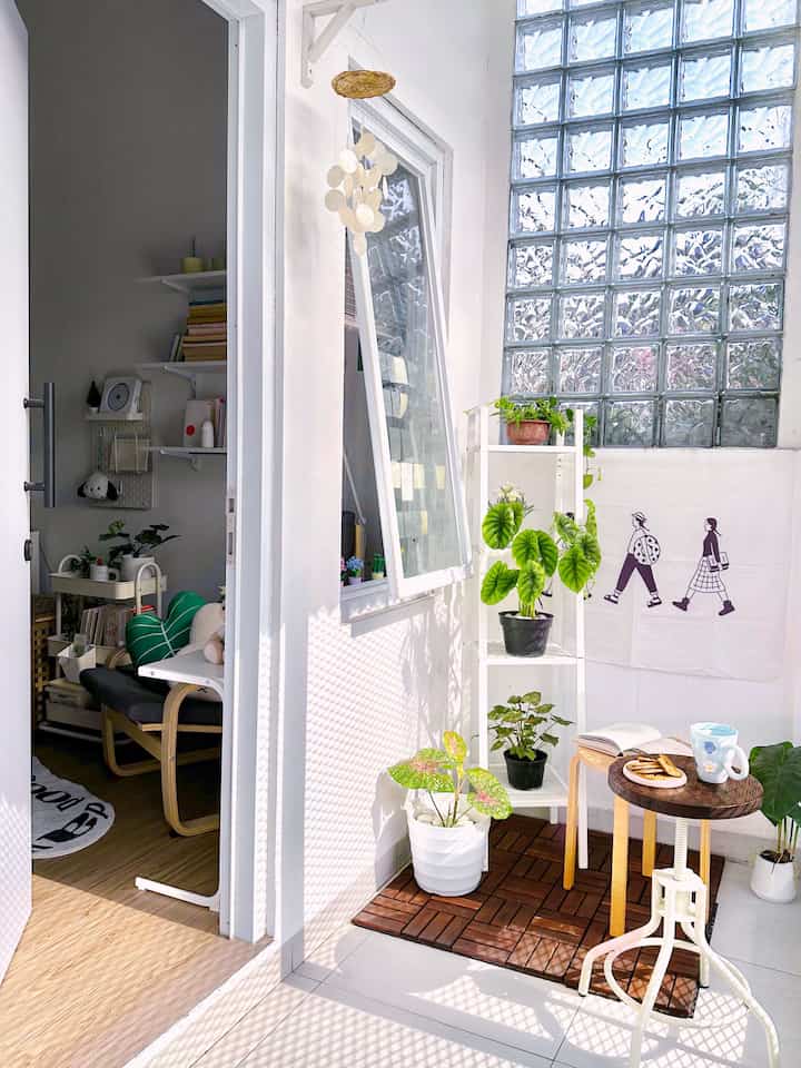 A bright white and wood-tone minimal balcony featuring green plants and a small stool table, creating a cozy atmosphere