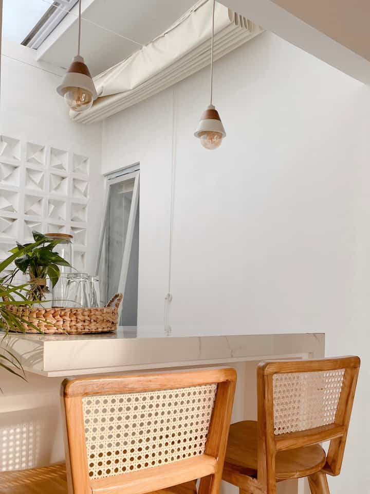 sebuah sudut dapur atau bar dengan desain minimalis dengan ukuran meja bar 100 x 50 ini sudah cukup untuk keluarga kami yang masih ber 3 .

- Kursi Bar : Terdapat dua kursi bar berbahan kayu dengan sandaran anyaman rotan, memberikan sentuhan alami pada ruangan. Desainnya yang sederhana namun elegan cocok dengan tema minimalis.

- Meja bar memiliki permukaan putih mengkilap, terbuat dari granit . Bagian tepi meja memiliki desain yang bersih dengan sudut yang tajam.

- Lampu gantung : Terdapat dua lampu gantung dengan kap berbentuk kerucut yang sederhana, dengan warna putih dan detail kayu. Lampu ini menambah suasana hangat dan nyaman dalam ruangan.

- Aksesoris Dekoratif : Di atas meja terdapat keranjang anyaman yang berisi beberapa item, termasuk tanaman hijau kecil dan botol kaca, memberikan elemen dekoratif yang simpel namun menarik.

Ruangan ini didominasi oleh warna putih yang cerah, dengan pencahayaan alami
menciptakan suasana yang terang, bersih, dan nyaman.

#DiningRoomStyling
#InspirasiRuangMakan
#RuangMakanMinimalis
#dekorasiruangmakan
#minimalis 
#StylingRuangMakan
