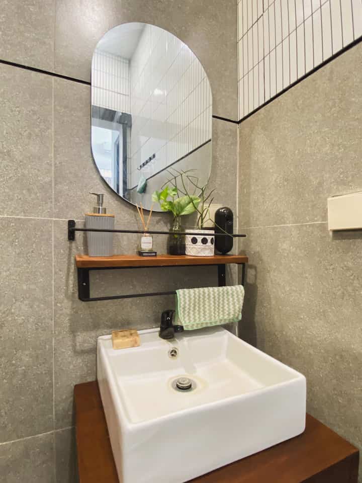 Bathroom washbasin space with gray tiles and wood-tone shelf, featuring an oval mirror and diffuser for a clean atmosphere