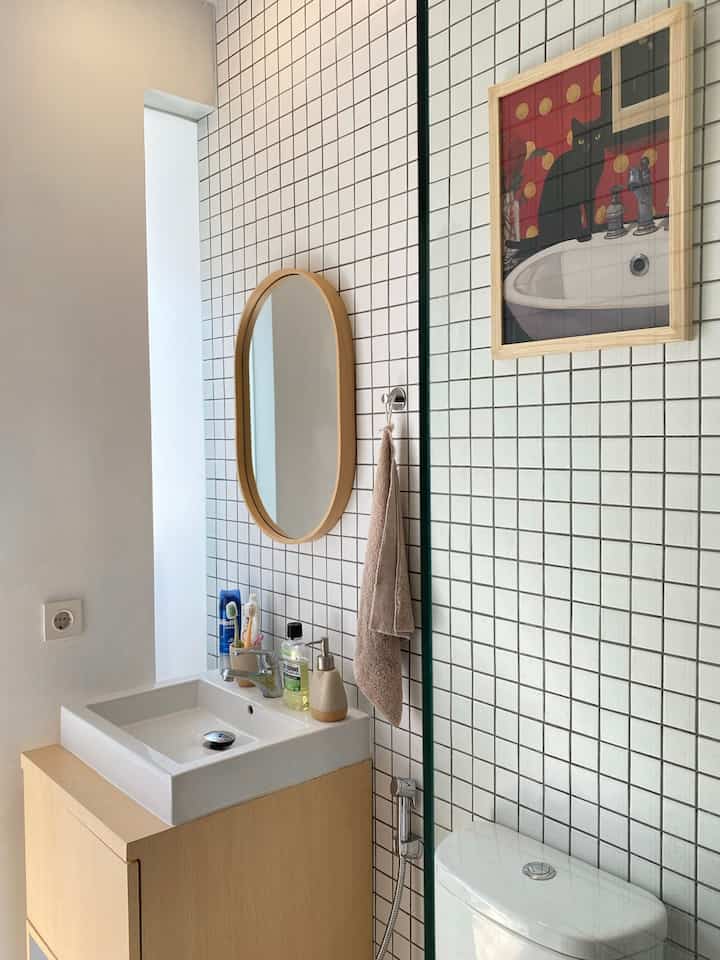 White and wood tone bathroom with tiled wall, oval mirror, sink, and toilet arranged in a clean design space