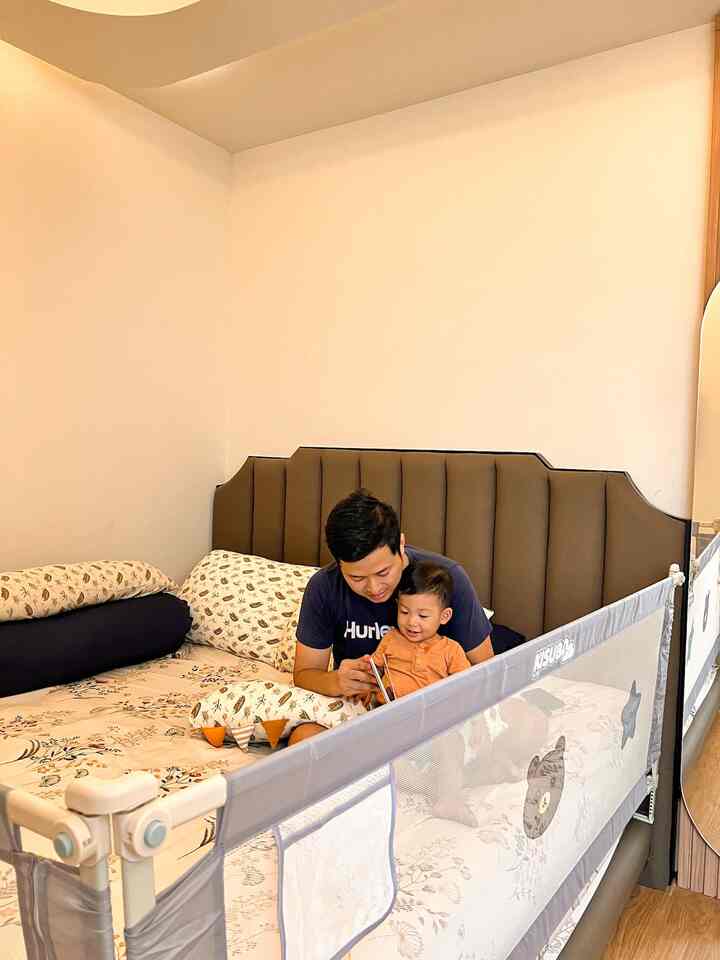 Beige-walled bedroom with brown bed headboard, featuring a man and baby reading on the bed with a protective safety rail and warm atmosphere
