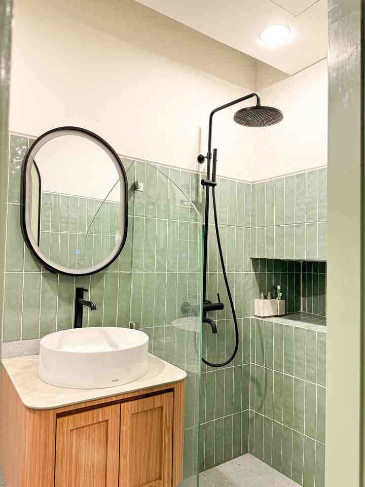 Natural green and white toned bathroom featuring a round mirror, black shower, and wooden vanity with a white basin in a clean setting