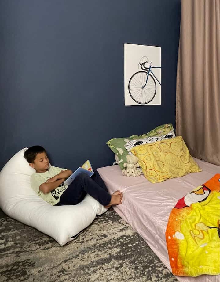 Sudut kamar ini adalah definisi kenyamanan sederhana yang tetap penuh gaya dan ramah anak. Dinding biru tua memberikan kesan tenang sekaligus modern, kontras dengan aksen warna-warni ceria pada kasur yang menambah semangat di ruang ini. Bean bag putih yang empuk menjadi spot favorit si kecil untuk membaca buku sebelum tidur, menciptakan sudut baca yang nyaman dan menyenangkan. Lukisan sepeda di dinding menambah sentuhan playful, membuat kamar ini terasa lebih hidup dan seru. Tirai berwarna netral serta karpet motif abstrak menyempurnakan kesan cozy, sambil tetap mempertahankan keamanan dan kenyamanan untuk anak. 

Dengan penataan yang fungsional dan estetis, ruang ini menunjukkan bagaimana rumah bisa tetap stylish dan ramah anak tanpa mengorbankan keamanan dan kenyamanan. 🌟 #InspirasiKamarAnak #HomeDecorAnak #RumahRamahAnak #CozyVibesKidsBedroom 