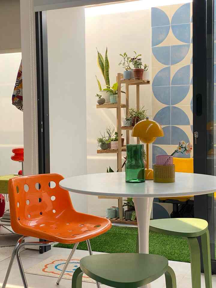 Bright natural balcony space featuring a white round dining table, an orange chair, and wooden rack with plants creating cozy home cafe atmosphere
