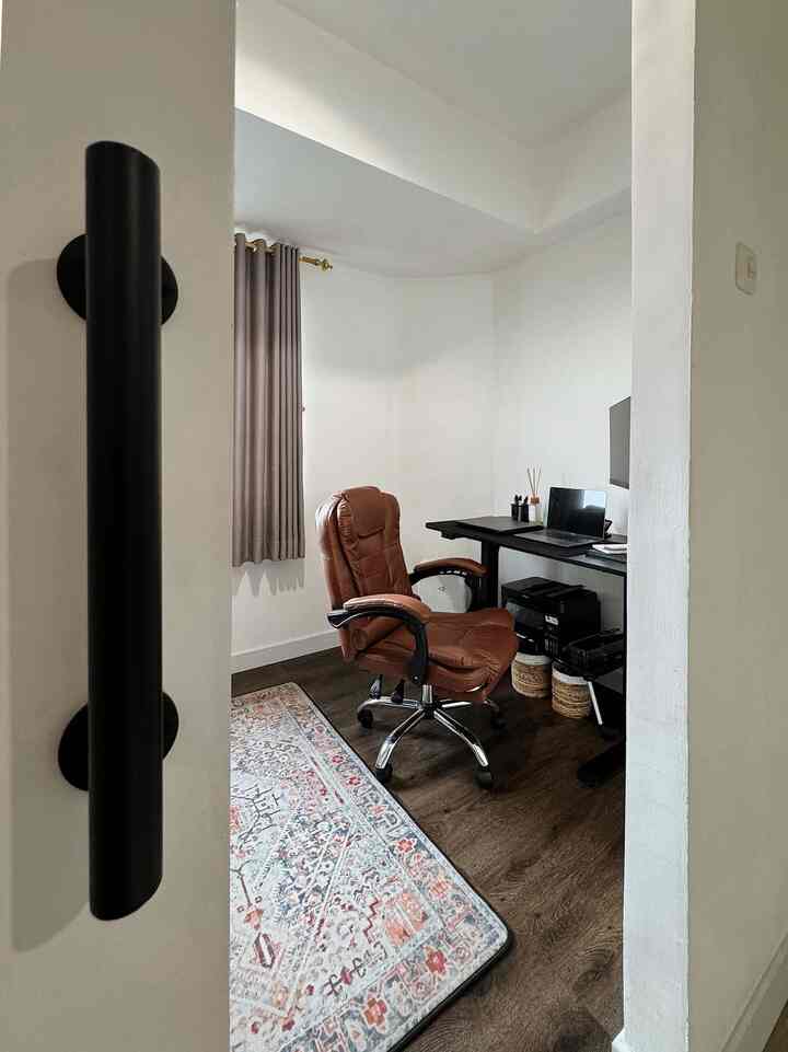 Compact home office with white walls and dark brown floor, featuring a brown office chair and black desk arranged for work