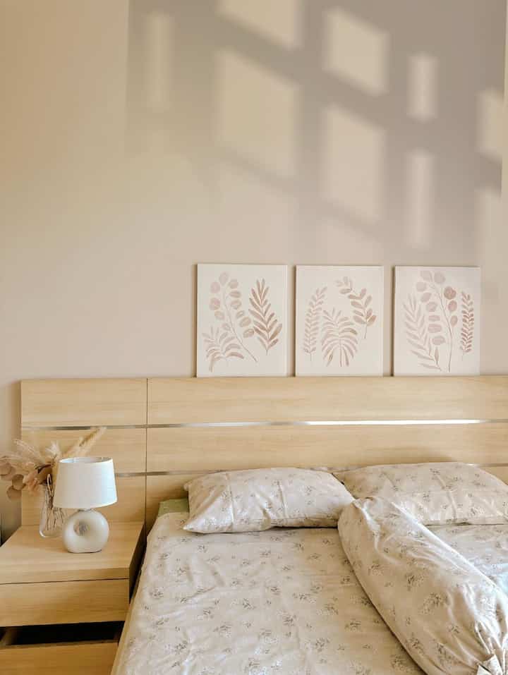 Beige-toned bedroom featuring wooden bed, bedding, nightstand with table lamp, plants, and botanical wall art creating a cozy atmosphere
