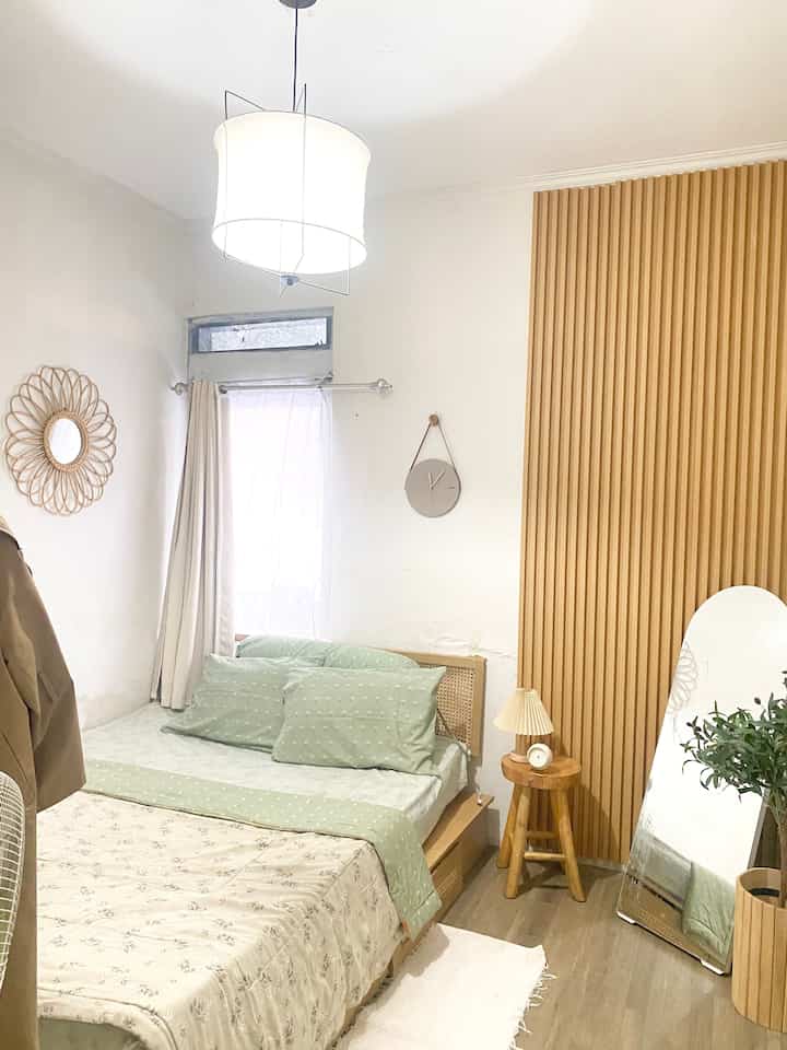 Compact bedroom with clean white walls and wood-tone wallpanel, featuring a wooden stool, standing mirror, and artificial olive tree creating cozy atmosphere