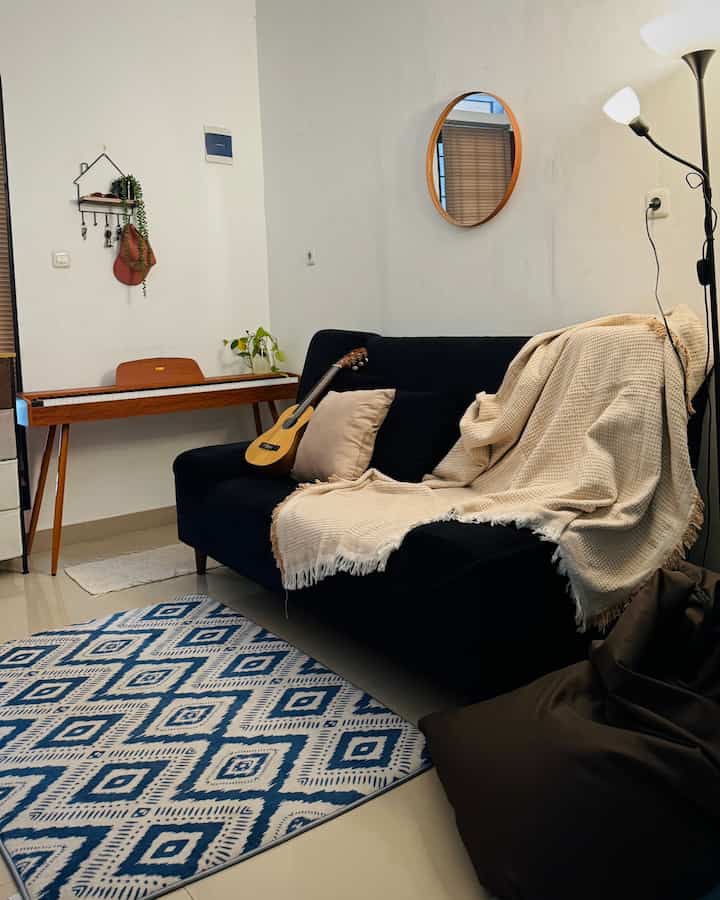 Natural-toned living room featuring a black sofa with beige throw, blue and white patterned carpet, and floor lamp on the right, creating a cozy family space