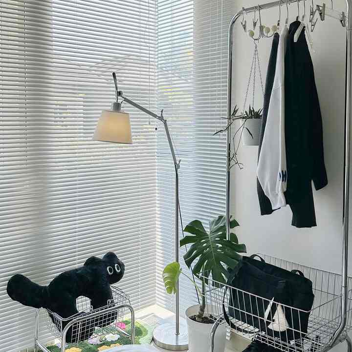 Simple and clean bedroom corner with white blinds and black cushion, featuring floor lamp and plants as focal points