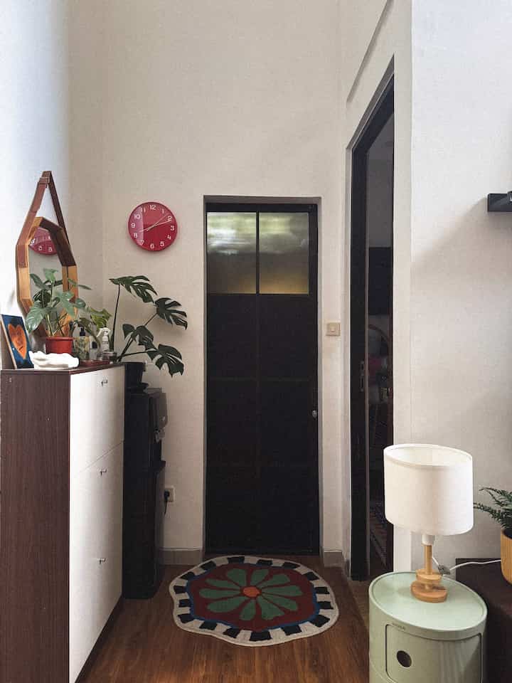 White and light brown toned entrance featuring shoe cabinet, green plant, and red wall clock with a simple atmosphere