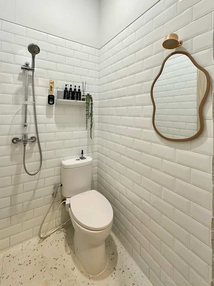 Modern small bathroom with white tiled walls, terrazzo floor, wooden framed mirror, shower head, and a small storage shelf
