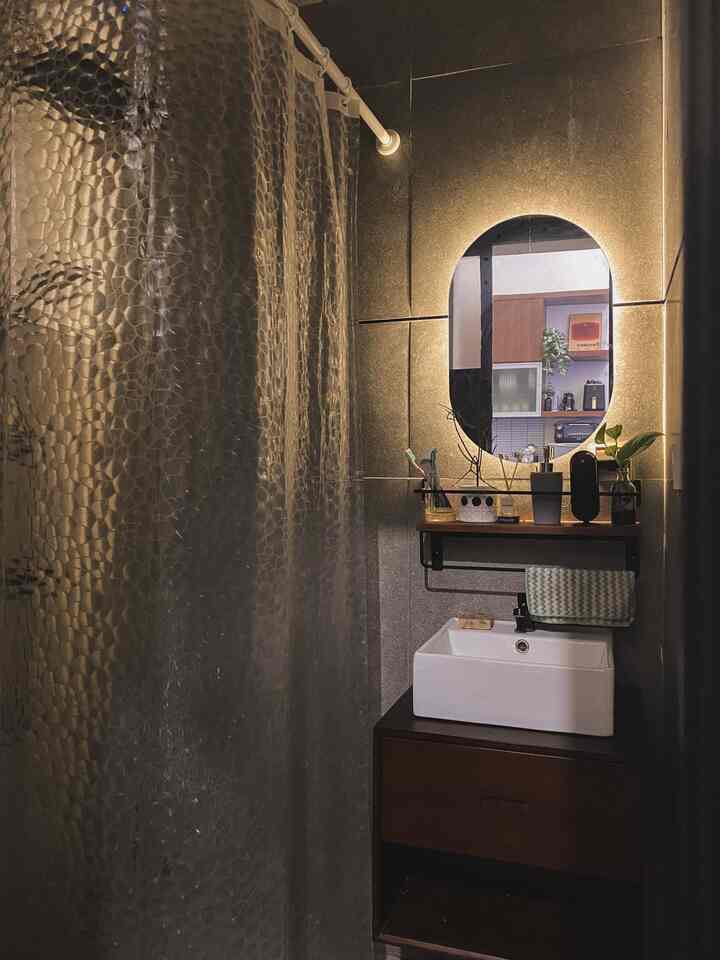 Dark black tiled narrow vertical bathroom featuring brown cabinet, backlit oval mirror, and textured shower curtain