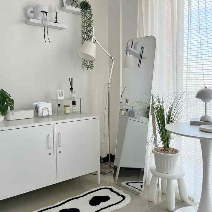Bright white-toned bedroom featuring a storage cabinet on the left, a large standing mirror, floor lamp, and green plants creating a clean, minimal space