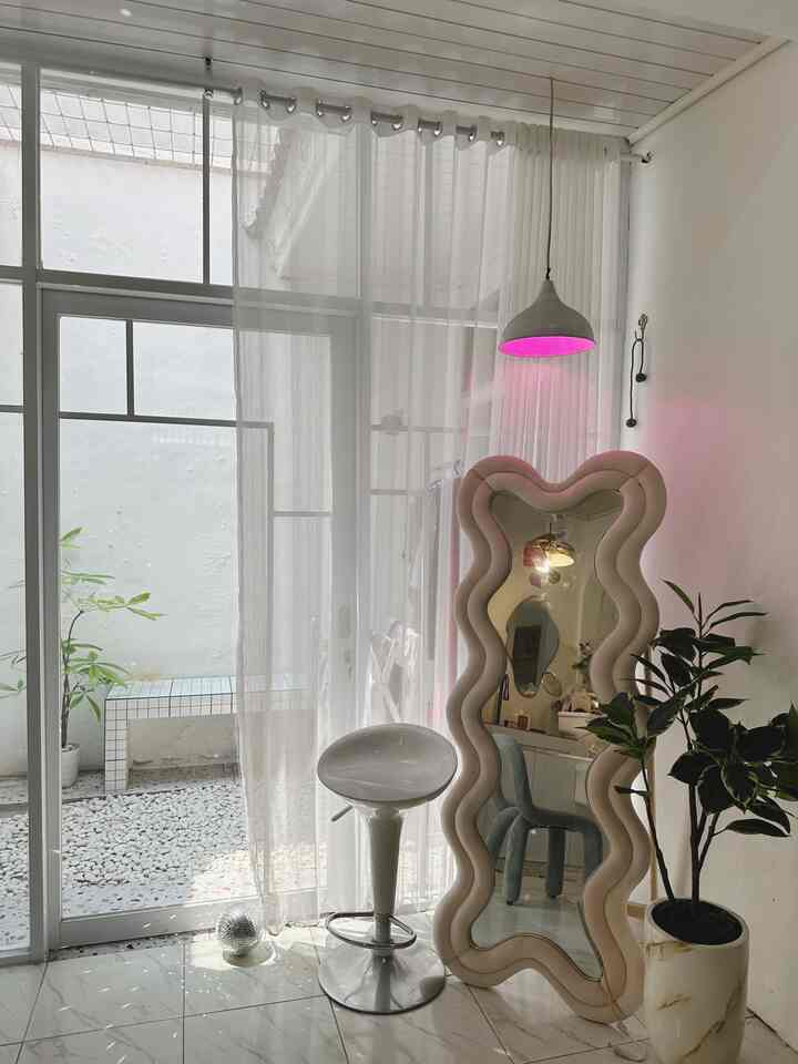 Modern corner space in white tones featuring a curvy standing mirror, bar stool, transparent curtains, and a potted plant with clean aesthetic