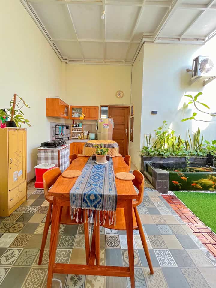 Beige walls with wood-tone dining table and chairs in dining and kitchen area, semi-outdoor with fish pond and plants on right