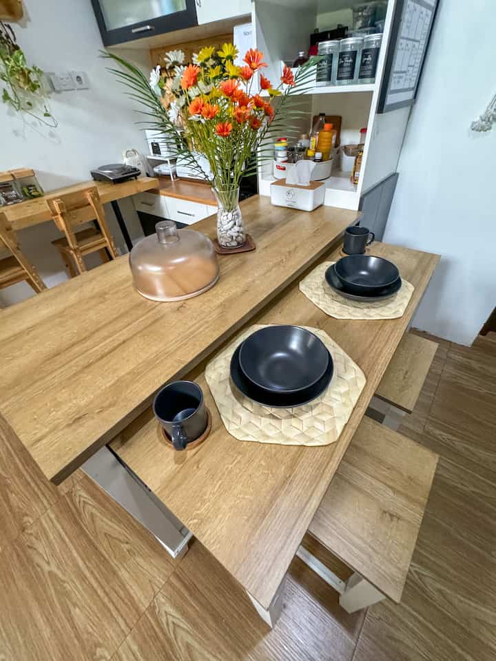 Wood tone kitchen dining space featuring a wooden dining table and benches with navy tableware and a vase of artificial flowers creating a neat atmosphere