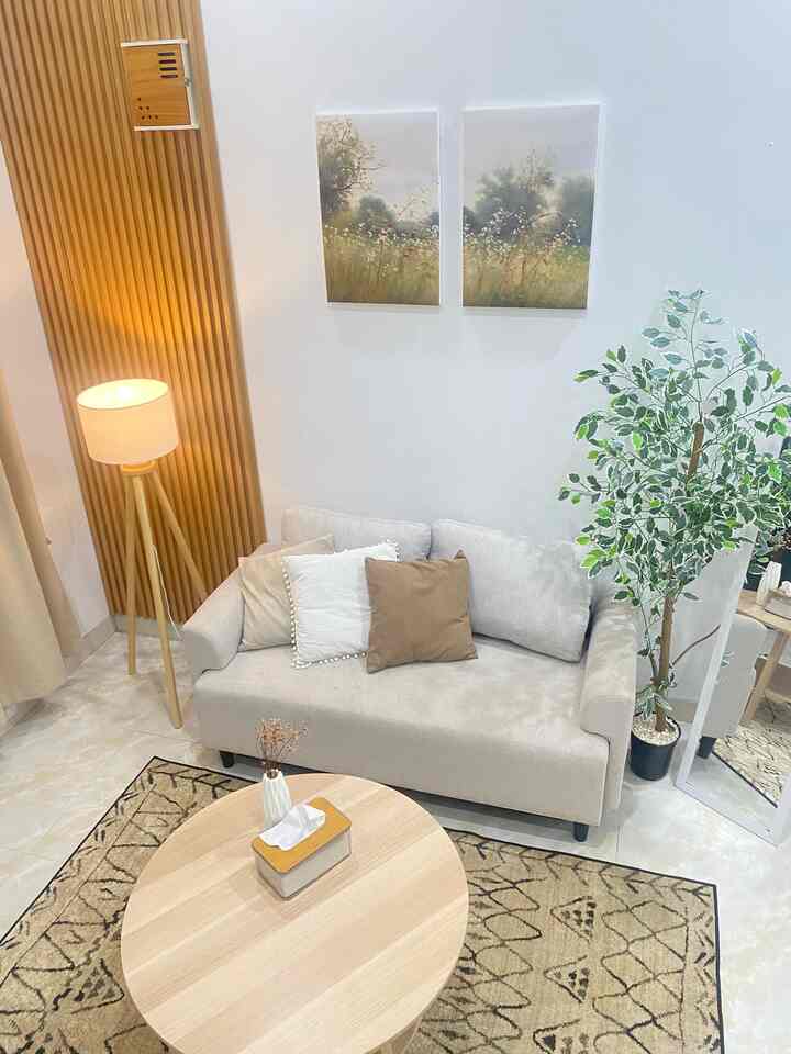 Natural toned living room featuring beige sofa, wooden round table, floor lamp, potted plant, and wall decor in a clean setting