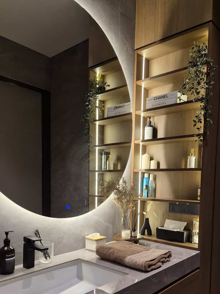 Kamar mandi ini menampilkan desain modern dan elegan, dengan kombinasi material yang mewah dan fungsional. Dinding dan lantainya menggunakan granite tile abu-abu yang memberikan kesan bersih dan berkelas, serta menambah dimensi visual dengan tekstur alami. Top table di area wastafel juga menggunakan granite tile, memberikan kesan solid dan tahan lama. Area shower dipisahkan dengan sekat kaca transparan, membuat ruangan terlihat lebih luas dan menjaga kesan minimalis.

Kabinet wastafel terbuat dari AICA HPL, material berkualitas tinggi yang tahan lama dan menambah sentuhan estetis pada kamar mandi dengan warna cokelat tua yang hangat. Selain memberi kesan modern, kabinet ini juga memiliki ruang penyimpanan tersembunyi, sehingga area wastafel tetap terlihat rapi.

Sanitair yang digunakan di kamar mandi ini adalah dari merek Kohler dan Toto, yang dikenal dengan kualitas dan desainnya yang elegan. Cermin bulat besar dengan lampu LED di belakangnya menambah pencahayaan lembut yang menciptakan suasana cozy dan stylish, sedangkan pencahayaan di bawah kabinet memberikan efek melayang yang menarik, sekaligus menciptakan atmosfer yang nyaman dan intim.

Perpaduan material berkualitas tinggi dan pencahayaan modern ini menjadikan kamar mandi terasa nyaman, fungsional, dan penuh gaya—cocok untuk generasi muda yang mengutamakan estetika dan kenyamanan dalam desain ruang pribadi mereka.

#KeramikKamarMandi 
#KlosetKamarMandi #tempattissue #wastafelkamarmandi #vase #cermin #handuk #tempatsikatgigi #lilinkamarmandi #bukupalsu  #WarnaKamarMandi 
#DindingKamarMandi #KeramikDinding #KeramikLantai #KamarMandiMinimalis #KamarMandiModern #KamarMandiGelap #InspirasiKamarMandi