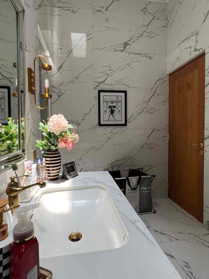 White marble tile bathroom with wood tone door in French Country style, featuring a vase with flowers and gold faucet at the washbasin