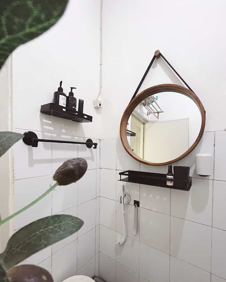 White tiled bathroom corner featuring a brown round mirror and black corner shelves