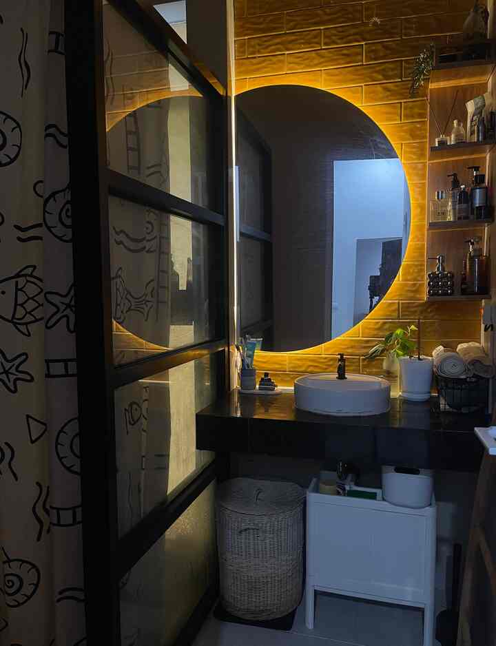 Bathroom with warm yellow lighting and round mirror, featuring a sink and organized storage shelves creating a cozy ambiance