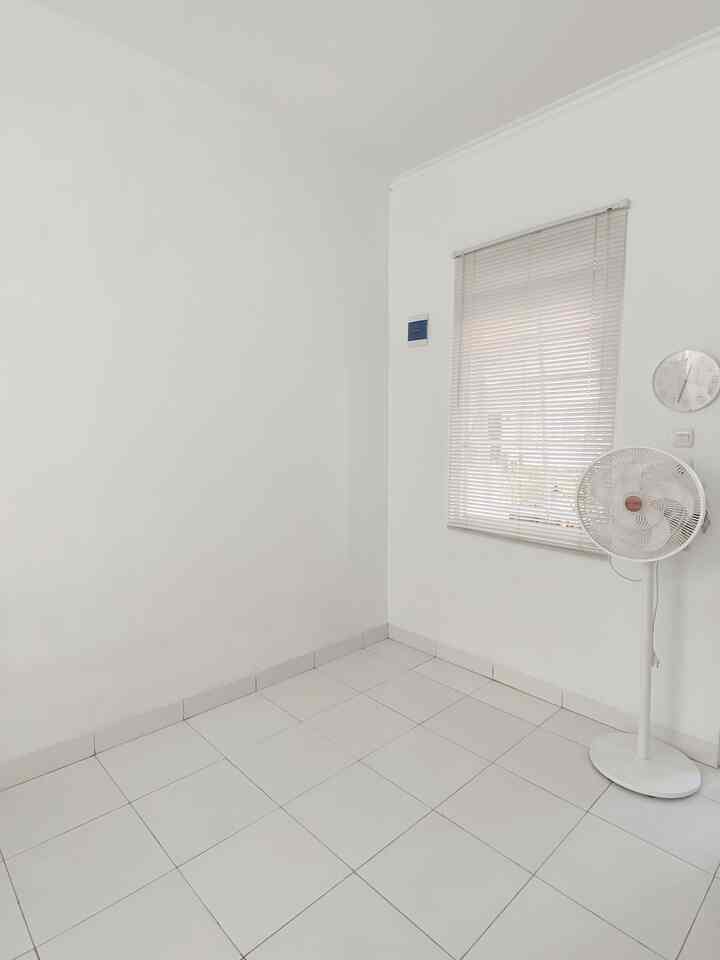 Small white-toned room with white walls and tiled floor, a window with blinds and a standing fan positioned on the right, featuring a minimalistic space