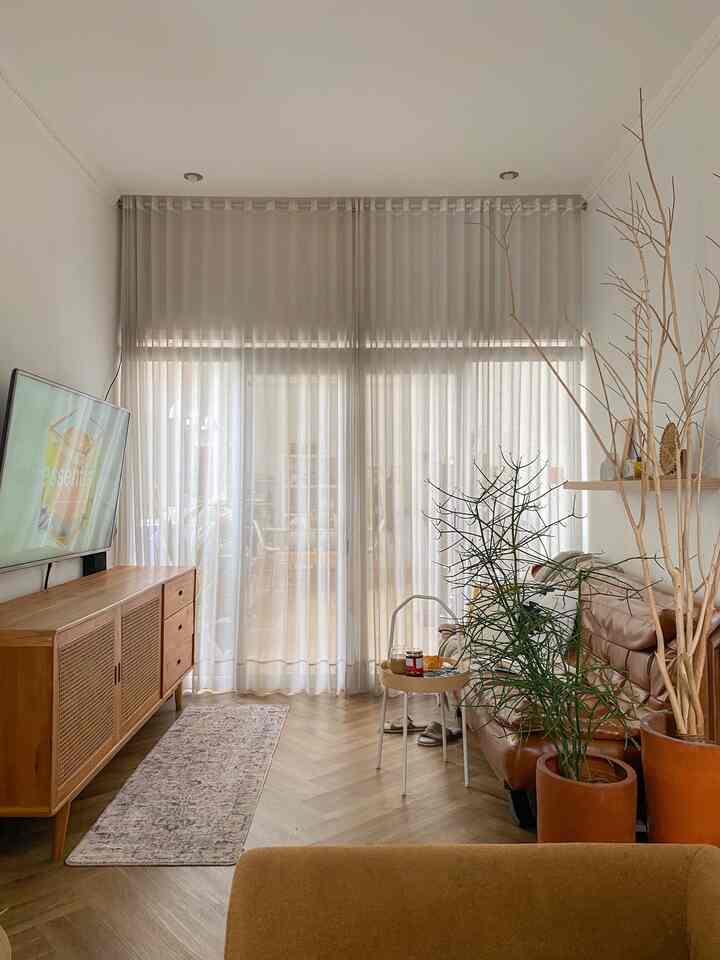 Natural-toned living room featuring brown leather sofa, wooden TV stand, and beige sheer curtains with a cozy atmosphere